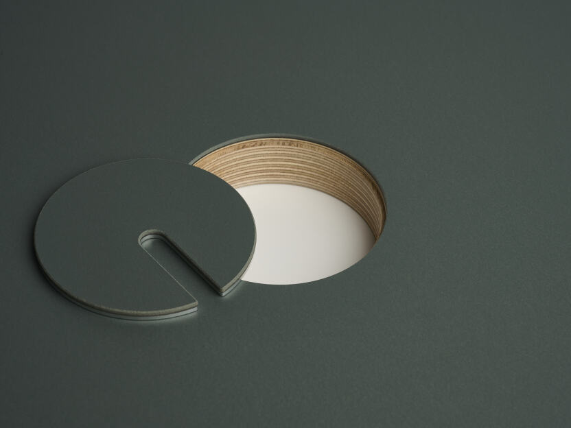 An anodised aluminum round cable lid by Daniel Lorch, covered with Conifer linoleum on one side. It sits next to its tabletop cut-out hole, revealing the plywood tabletop core material.