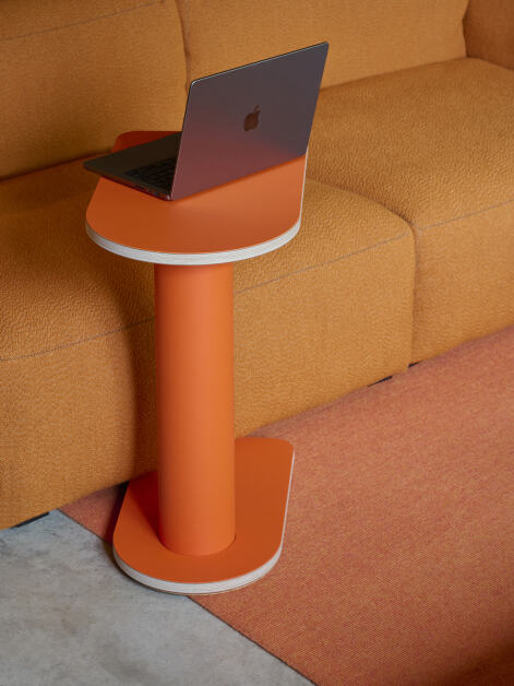 Ein halbrunder Off-Cut Beistelltisch mit einer Tischplatte aus Sperrholz, bedeckt mit orangefarbenem Linoleum, wird als Arbeitsplatz für einen Laptop neben einem Sofa in einem Wohnzimmer oder Gemeinschaftsbüro verwendet.