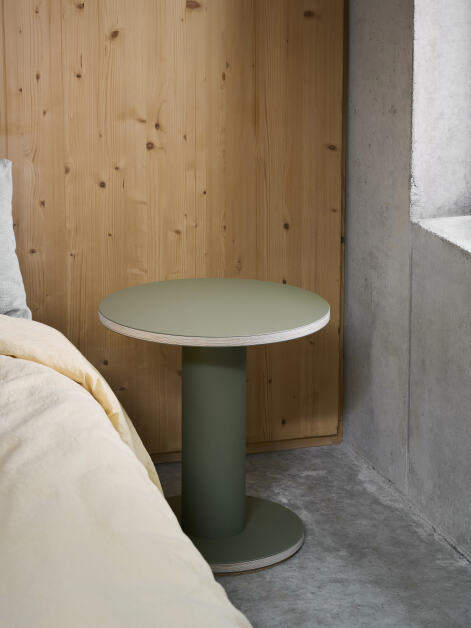 Round Off-cut side table with a plywood tabletop covered with olive linoleum, suitable for use as a bedside table in a bedroom in a private home or hotel.