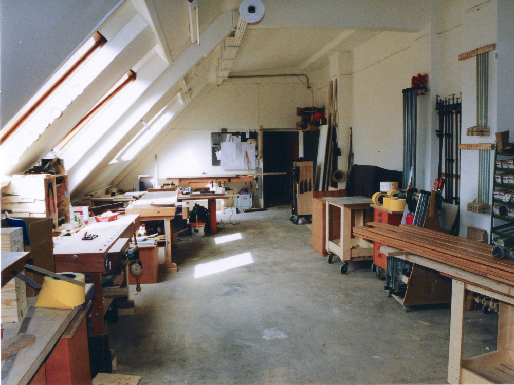 Blick in die erste Werkstatt in Berlin mit Werkbänken, Holzmaterialien und Werkzeugen unter einem Dachgeschossfenster.