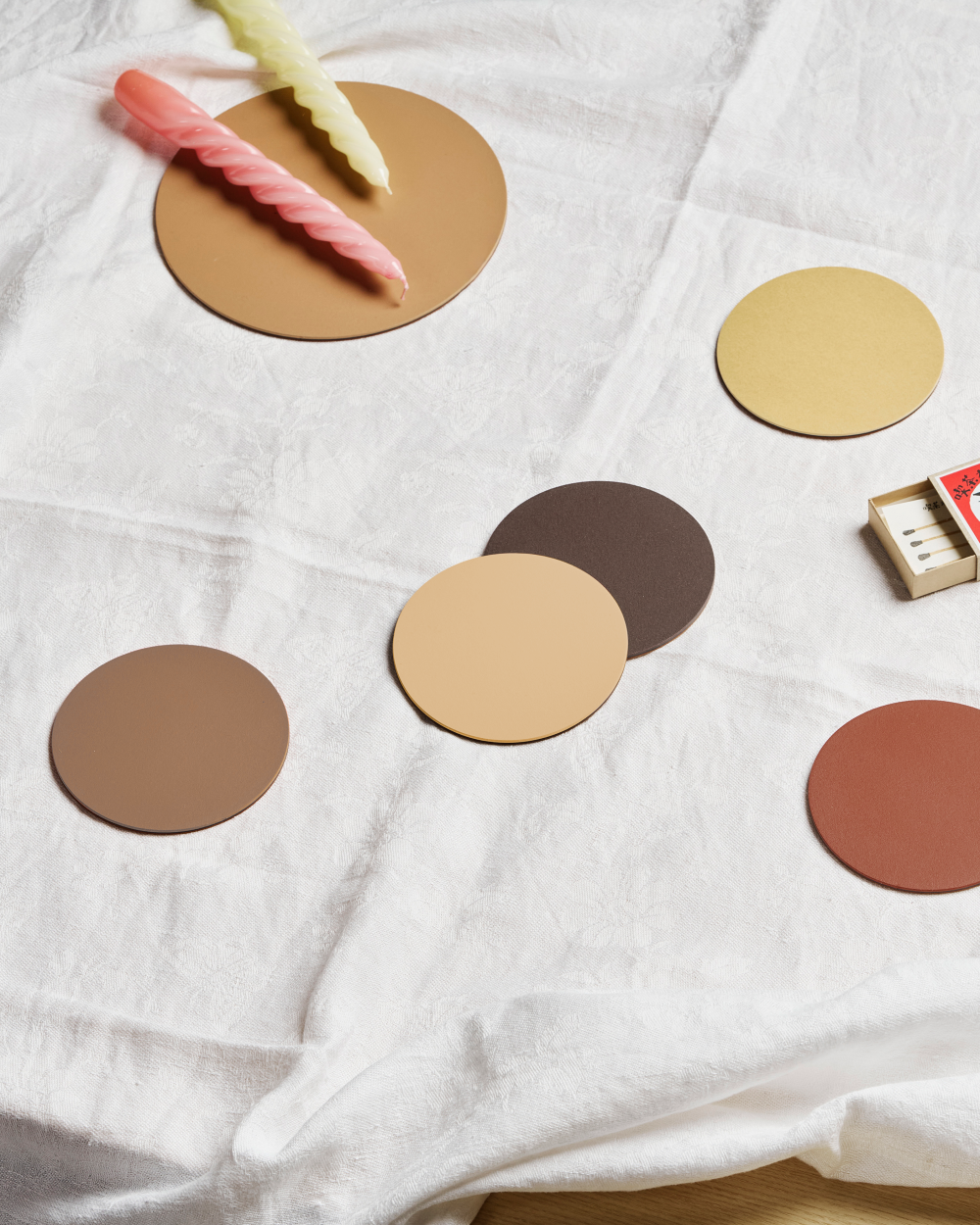 PAD Untersetzer aus Linoleum in warmen Farbtönen auf weißem Tischtuch arrangiert, mit farbigen Kerzen und Streichholzschachtel.