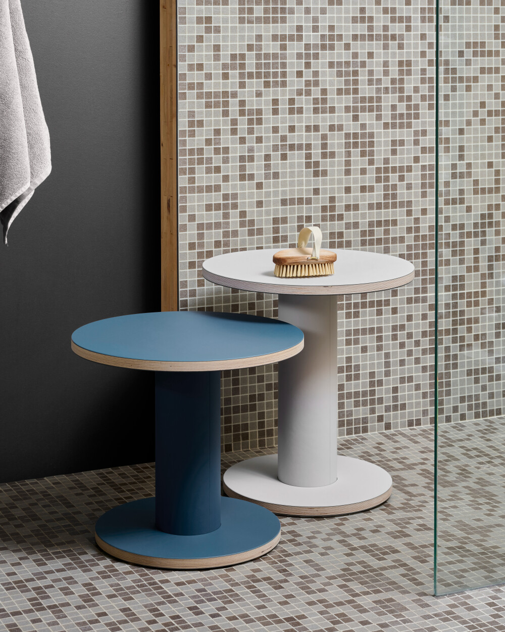 Two round OFF-CUT side tables by Faust with linoleum surfaces, in jeans blue and light gray, placed in a tiled bathroom.