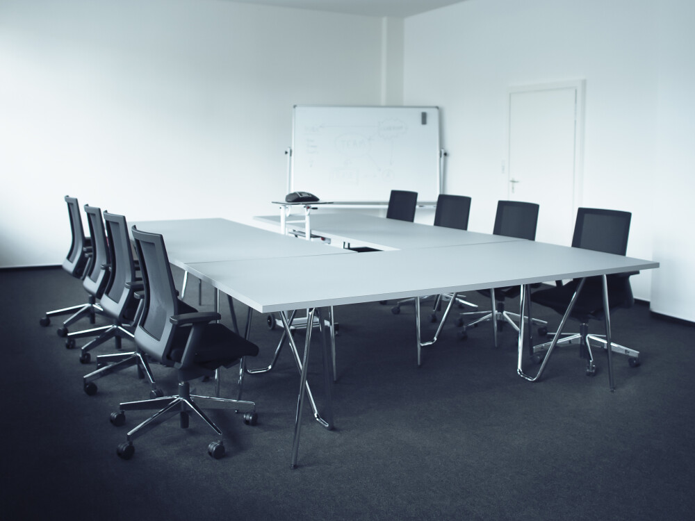 Grey Conference table with multiple panels on chrome Sinus table trestles, surrounded by black office chairs.