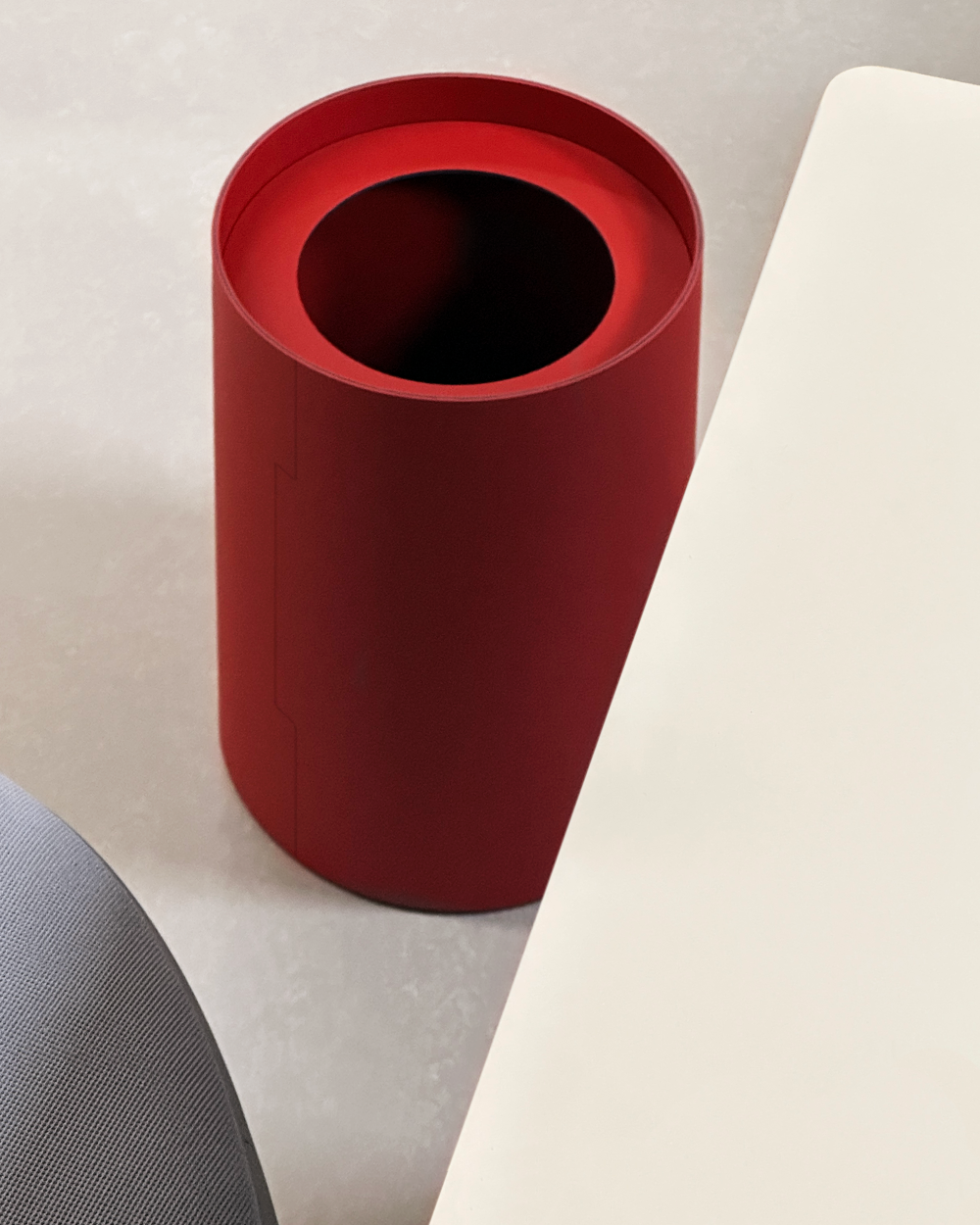 Cylindrical TIDY wastepaper basket made of red linoleum on a light-coloured floor, next to a white table and grey chair.