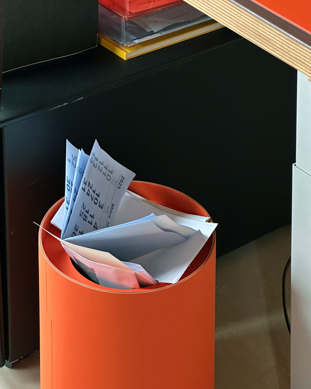 Cylindrical TIDY wastepaper basket made of orange linoleum, filled with paper waste, next to a desk.