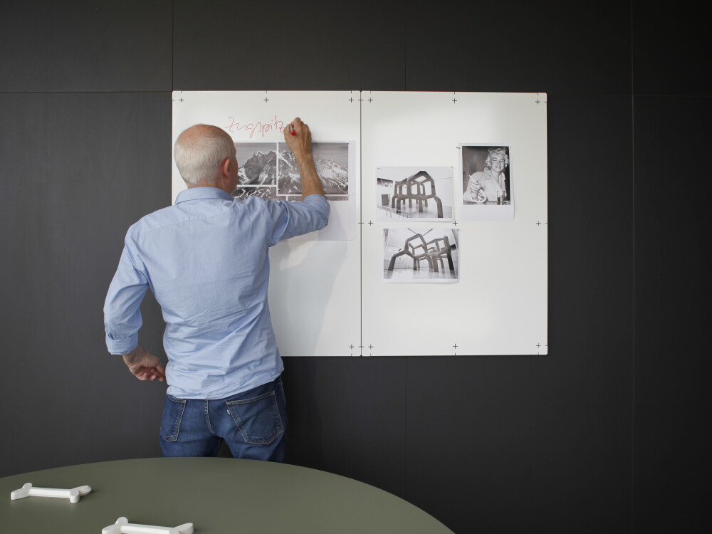 White magnetic boards on a dark wall, with pinned photographs and a person writing on them with a marker pen.